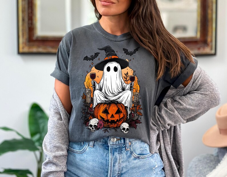 May include: Dark gray t-shirt featuring a Halloween-themed graphic. The design includes a ghost wearing a witch hat, sitting on a carved pumpkin, holding wine glasses, with bats and skulls. The shirt is made of a soft material.