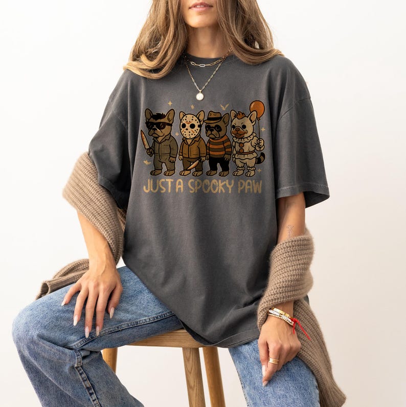 May include: Dark gray t-shirt featuring a graphic of four French bulldogs dressed as horror movie characters. The phrase "JUST A SPOOKY PAW" is printed below the dogs in a gold color. The shirt is a relaxed fit.