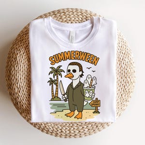 May include: White t-shirt featuring a cartoon duck dressed as a horror movie character, holding a knife and a bucket of ghosts. The shirt has the text "SUMMERWEEN" above the graphic and "CAMP SPOOKY" on a sign. The design includes palm trees and a beach scene.