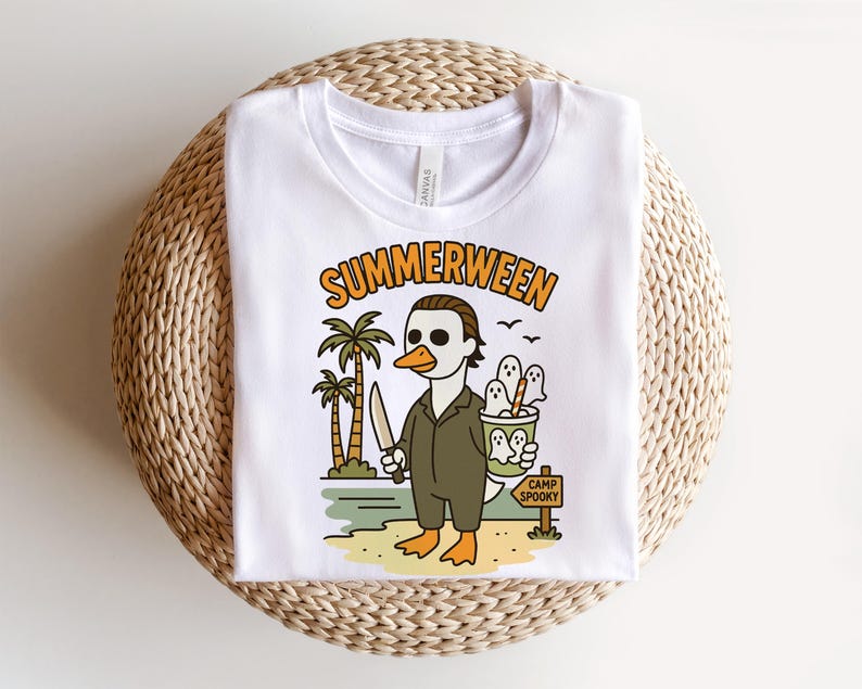 May include: White t-shirt featuring a cartoon duck dressed as a horror movie character, holding a knife and a bucket of ghosts. The shirt has the text "SUMMERWEEN" above the graphic and "CAMP SPOOKY" on a sign. The design includes palm trees and a beach scene.