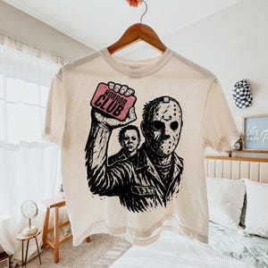 Pu&ograve; includere: T-shirt color crema con grafica di Jason Voorhees di Venerd&igrave; 13 e Michael Myers di Halloween. Il design include il testo "HORROR CLUB" su un rettangolo rosa. La maglietta &egrave; appesa a un appendiabiti di legno.