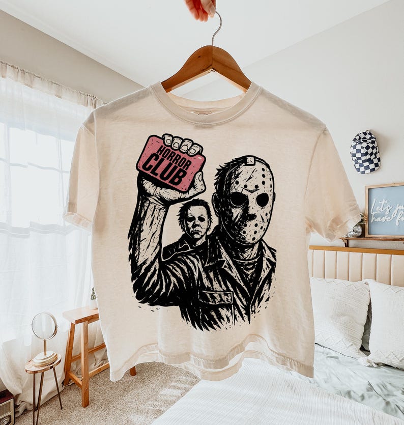 Pu&ograve; includere: T-shirt color crema con grafica di Jason Voorhees di Venerd&igrave; 13 e Michael Myers di Halloween. Il design include il testo "HORROR CLUB" su un rettangolo rosa. La maglietta &egrave; appesa a un appendiabiti di legno.
