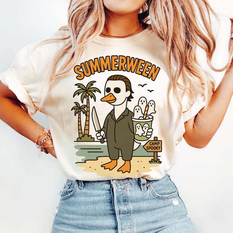 May include: Cream-colored t-shirt with the word "SUMMERWEEN" in orange lettering. The graphic features a duck dressed as a horror movie character holding a knife, with ghosts in a bucket and palm trees. The sign reads "CAMP SPOOKY".