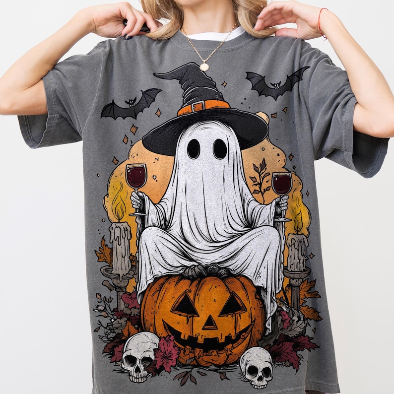 May include: Gray t-shirt featuring a Halloween-themed graphic. The design includes a ghost wearing a witch hat, holding wine glasses, and sitting on a jack-o'-lantern. Bats, candles, and skulls complete the spooky scene.