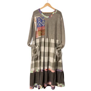 May include: A long-sleeved dress with a patchwork design on the upper left chest. The dress features a gray top, a plaid midsection, and a dark gray ruffled hem. It has two pockets and is hanging on a wooden hanger.