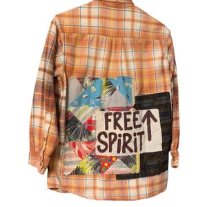 May include: Orange plaid button-down shirt with patchwork details. The back features a colorful patchwork design and the words "FREE SPIRIT" with an upward arrow. The shirt has long sleeves and buttoned cuffs.