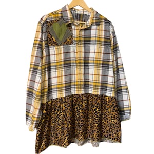 May include: A long-sleeve button-down shirt featuring a yellow, white, and gray plaid pattern. The shirt includes a chest pocket with a green heart-shaped patch over a leopard print square, and a gathered leopard print skirted bottom hem.