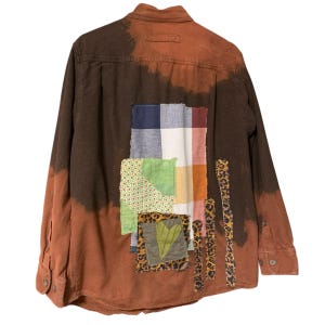 May include: A long-sleeve button-down shirt featuring a brown and rust-colored tie-dye effect. The back is decorated with a large rectangular plaid patch, a green patterned square, a leopard print strip, and a heart-shaped fabric applique.