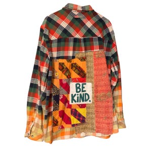 May include: A long-sleeve button-down shirt featuring a red, green, and orange plaid pattern. The back displays a large, multi-colored patchwork panel with various fabric textures and a white rectangular patch with the text BE KIND.