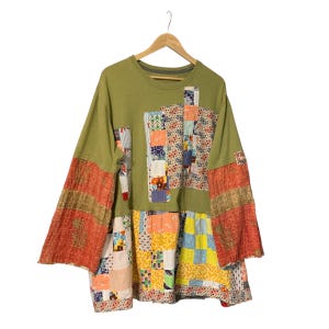 May include: A long-sleeve olive green shirt featuring a patchwork design. The front panel and sleeves are adorned with various colorful fabric squares in floral, geometric, and polka dot patterns. The lower section is a wide, multi-colored quilt panel.