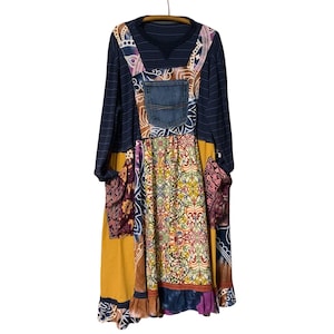 May include: A long-sleeved patchwork dress with a denim pocket and a variety of patterned fabrics. The dress features a navy blue striped top, a mustard yellow skirt, and pockets with floral and paisley designs. It hangs from a wooden hanger.