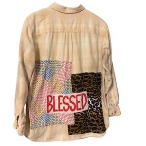 May include: Tan plaid button-down shirt with patchwork details. The back features a patchwork design with the word "BLESSED" in red letters, along with a leopard print and geometric patterns. The shirt has long sleeves and a collared neckline.
