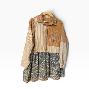 May include: A long-sleeve button-down shirt featuring a patchwork design. The upper section combines tan and white vertical stripes with a brown plaid pattern and a chest pocket. The lower section is a blue fabric with a small yellow floral print.