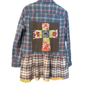 May include: Blue plaid button-down shirt with a patchwork cross design on the back. The cross is made of various floral and patterned fabrics. The bottom of the shirt has a ruffled hem with a red, white, and blue plaid pattern. The word "dee" is visible on the cross.