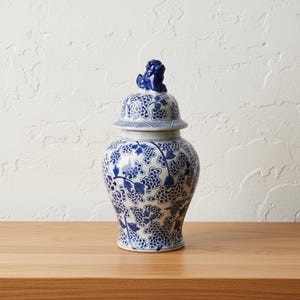 May include: A blue and white ceramic ginger jar with a lid. The jar has a floral pattern in blue on a white background. The lid is topped with a small blue figurine. The jar is on a wooden surface.