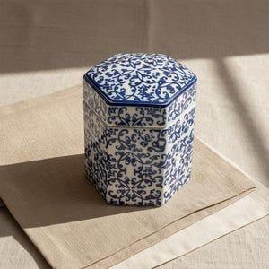 May include: A hexagonal ceramic box with a lid, decorated with a blue and white floral pattern. The box sits on a beige linen surface, with shadows from sunlight. The design is reminiscent of classic porcelain.