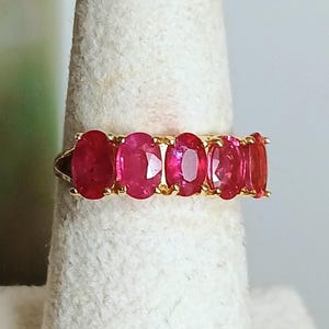 May include: A gold ring featuring five oval-cut rubies. The rubies are a vibrant red color and are set closely together in a row. The ring is displayed on a white surface, showcasing the gemstones and the gold band.