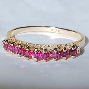 May include: A gold ring with a row of vibrant pink gemstones. The ring's band is a warm gold color, and the gemstones are set closely together, creating a continuous line of color. The ring is set on a white surface.