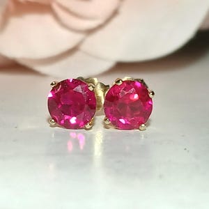 May include: A pair of stud earrings featuring round, faceted, vibrant pink-red stones set in four-prong gold metal settings. The stones exhibit a brilliant cut with clear light reflection, securely held by the metallic prongs on each earring base.
