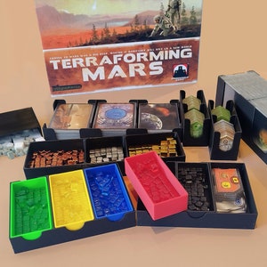 Pode incluir: Jogo de tabuleiro "Terraforming Mars" com vários componentes. A caixa do jogo é visível, juntamente com várias bandejas pretas cheias de peças de jogo coloridas, cartas e outros acessórios. Os componentes do jogo são organizados para jogar.
