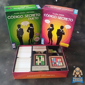 Può includere: Sono esposte due scatole di giochi da tavolo, "Código Secreto Dueto" e "Código Secreto". Le scatole sono verdi e rosa, con sagome di persone. All'interno, ci sono carte e tabelloni di gioco con quadrati e griglie colorate. Il gioco è di Vlaada Chvátil.