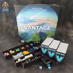 Op de afbeelding: Een bordspelset met de titel "VANTAGE" op de doos, met een landschapsillustratie. Zwarte opbergbakken zijn gevuld met spelstukken en kaarthouders met het "V"-logo en nummerreeksen zijn ook aanwezig.