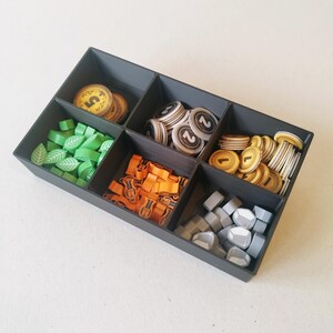 May include: A black rectangular plastic organizer tray with five compartments containing various game pieces. The sections hold green leaf-shaped tokens, orange figures, grey stone-shaped pieces, and circular tokens marked with the numbers 1, 2, and 5.