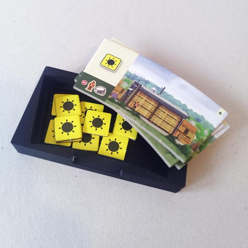 May include: A black rectangular plastic tray containing several square yellow tokens with black sun icons. Resting on top of the tray is a rectangular game card featuring an illustration of a building, a sun icon, a small figure, and a stone icon.