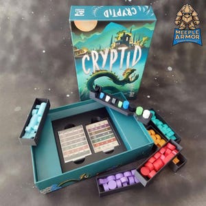 Könnte beinhalten: Brettspiel "Cryptid" mit einer grün-blauen Box. Die Box zeigt eine Landschaft mit einer Seeschlange. Im Inneren sind Spielsteine in Blau, Orange, Rot und Lila in Tabletts angeordnet.