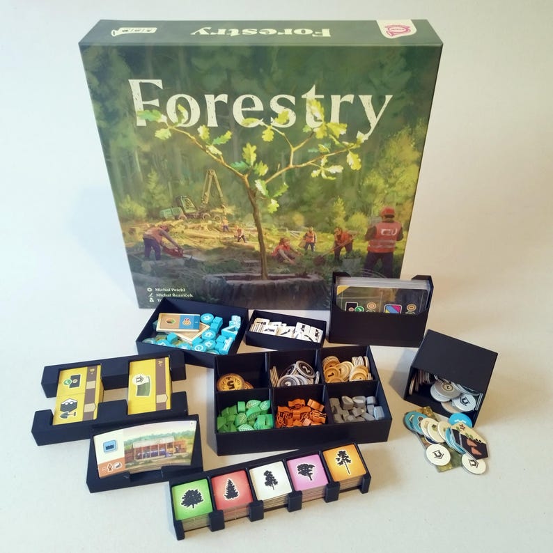 May include: A Forestry board game box featuring a forest illustration with workers, accompanied by black plastic organizers containing various game components including small wooden tokens, circular cardboard markers, and rectangular cards.