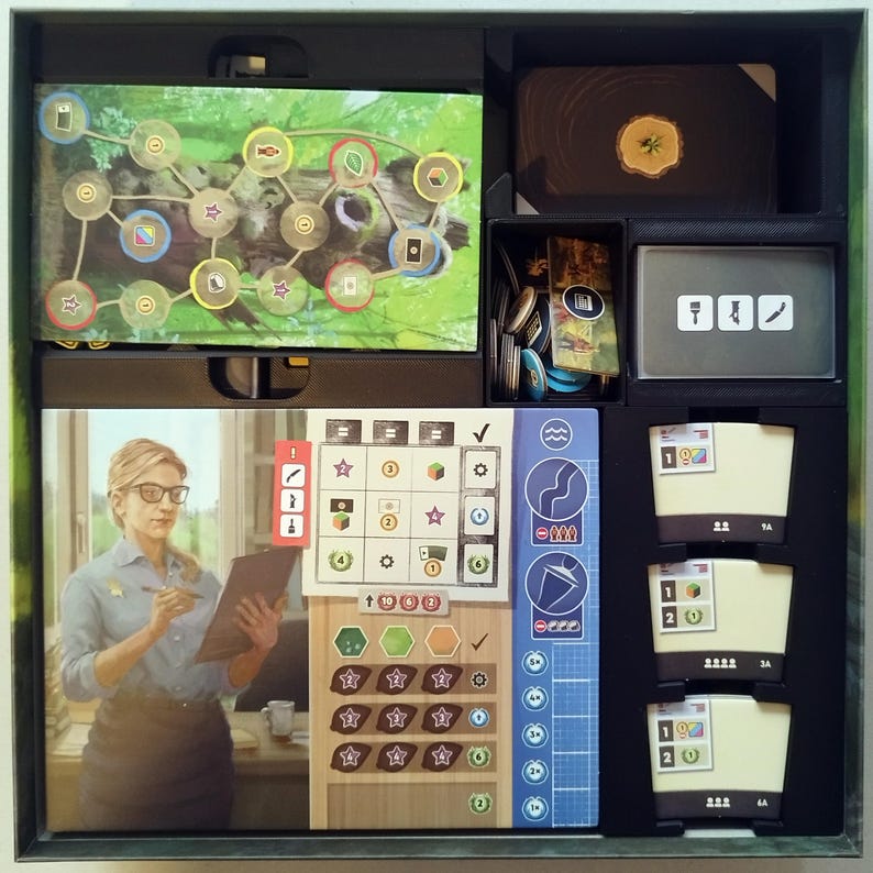 May include: A board game box interior containing a game board with a forest path illustration, a player board featuring an illustration of a person with glasses holding a clipboard, various card decks, and small circular tokens organized in black plastic trays.
