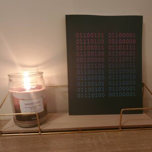 K&ouml;nnte beinhalten: Eine schwarze Karte mit einem Muster aus bin&auml;rem Code in Pink und Blau auf einem Holzregal mit einer brennenden Kerze in einem Glasgef&auml;&szlig;. Das Kerzenetikett lautet "Autumn Leaves" mit einem Design aus B&auml;umen und Bl&auml;ttern.