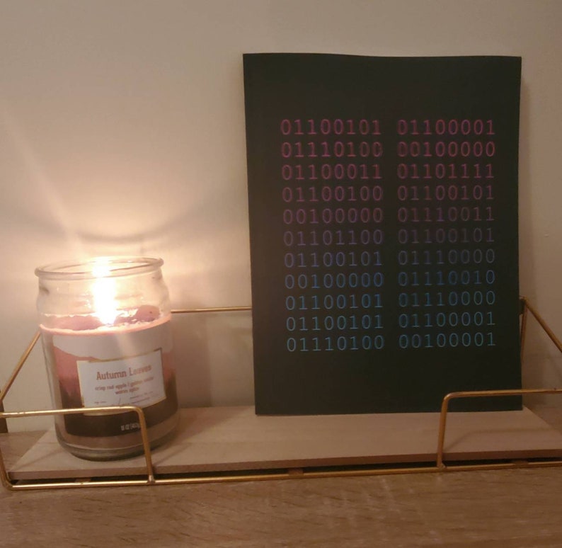 K&ouml;nnte beinhalten: Eine schwarze Karte mit einem Muster aus bin&auml;rem Code in Pink und Blau auf einem Holzregal mit einer brennenden Kerze in einem Glasgef&auml;&szlig;. Das Kerzenetikett lautet "Autumn Leaves" mit einem Design aus B&auml;umen und Bl&auml;ttern.