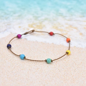 May include: A handmade bracelet featuring a series of colorful beads in a rainbow pattern. The beads are strung on a brown cord. The bracelet is displayed on a sandy beach with the ocean in the background.