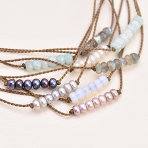 May include: A set of seven delicate necklaces with brown cord and various gemstone beads. The necklaces feature pearls, aquamarine, labradorite, and opal beads.