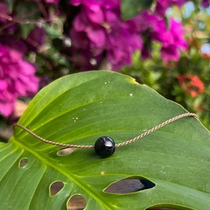 May include: A black bead necklace with a single black bead on a brown cord, resting on a green leaf.