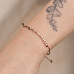 May include: A brown cord bracelet with a silver bamboo-shaped charm. The bracelet is adjustable and can be worn on the wrist.