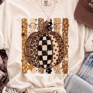 Op de afbeelding: Een ivoorkleurig t-shirt met een centrale pompoengrafiek met afwisselende segmenten van luipaardprint, zwart-wit geblokt en stippen. De pompoen is geplaatst tegen verticale penseelstreekstrepen en kleine stervormen.