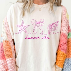 May include: Cream-colored t-shirt with a pink line drawing of a starfish, bow, and seashell, with the words "Summer vibes" in pink script. The person is wearing a colorful, patchwork cardigan.