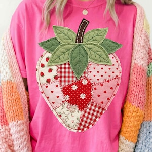 Könnte beinhalten: Ein pinkfarbenes T-Shirt mit einem großen Erdbeer-Design aus Patchwork. Die Erdbeere hat grüne Blätter, einen braunen Stiel und verschiedene gemusterte Flicken in Rot, Rosa und Weiß. Über dem T-Shirt wird eine farbenfrohe, gemusterte Strickjacke getragen.