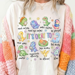 May include: Cream-colored t-shirt featuring colorful cartoon dinosaurs with phrases like "need routines" and "it's okay to." The shirt is paired with a multi-colored knitted cardigan. The design includes flowers and stars.