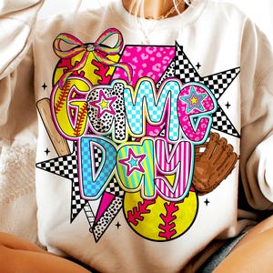 以下が含まれることがあります： 白地にカラフルなグラフィックデザインが施されたスウェットシャツ。"Game Day"の文字と、野球ボール、星、市松模様が描かれています。
