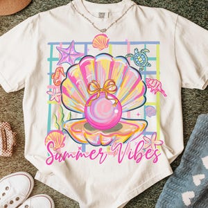 May include: Off-white t-shirt with a vibrant seashell graphic, including a pearl, starfish, and seahorse. The words "Summer Vibes" are written in pink script. A silver necklace and white trainers are also visible.