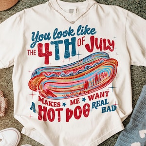 May include: Off-white t-shirt with a colorful graphic of a hot dog and text that reads "You look like the 4th of July makes me want a hot dog real bad." The design features red, blue, and white colors.