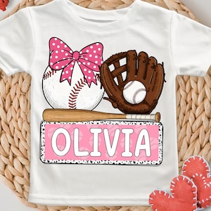 May include: White t-shirt with a pink bow, baseball, glove, and bat graphic. The text "OLIVIA" is on a pink banner with a black and white leopard print border.