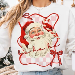 May include: White sweatshirt featuring a vintage illustration of Santa Claus with a red hat and beard, holding a candy cane. The design includes a red and pink checkered background and the words "ho ho ho".