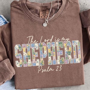 Pode incluir: Um moletom Comfort Colors marrom com o texto "The Lord is my Shepherd Psalm 23". A palavra "Shepherd" está em um design floral de patchwork. Um colar de prata está drapeado no moletom.