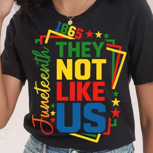Può includere: T-shirt nera con un design grafico colorato che celebra il Juneteenth. Il design include le parole "1865", "THEY NOT LIKE US" e "Juneteenth" in vari colori, insieme a stelle e forme geometriche.