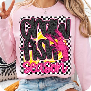 May include: Pink sweatshirt with a black and white checkered background. The text "Crawfish Season" is written in black with a glittery texture. Two pink crawfish are on a yellow lemon slice.