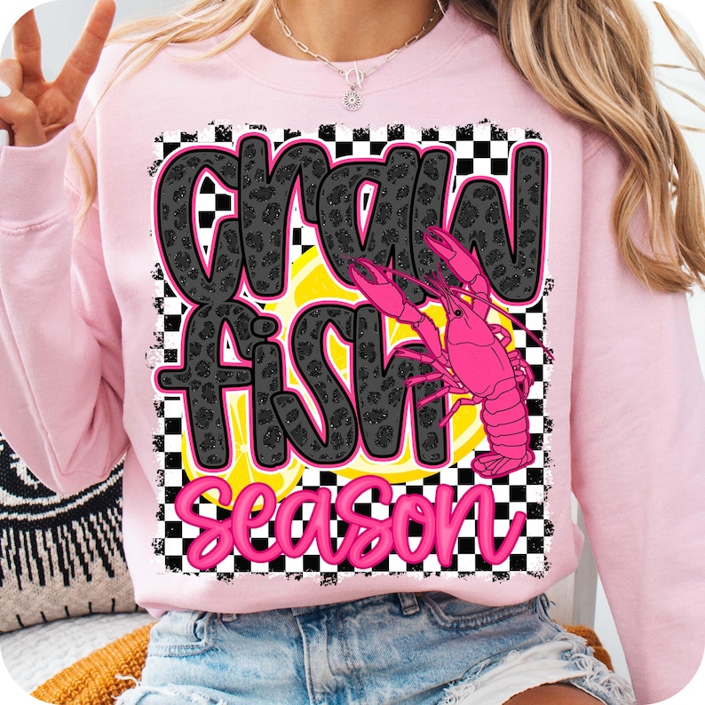May include: Pink sweatshirt with a black and white checkered background. The text "Crawfish Season" is written in black with a glittery texture. Two pink crawfish are on a yellow lemon slice.
